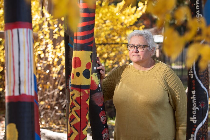A woman standing amongst Indigenous art, yellow leaves of trees surrounding her. 