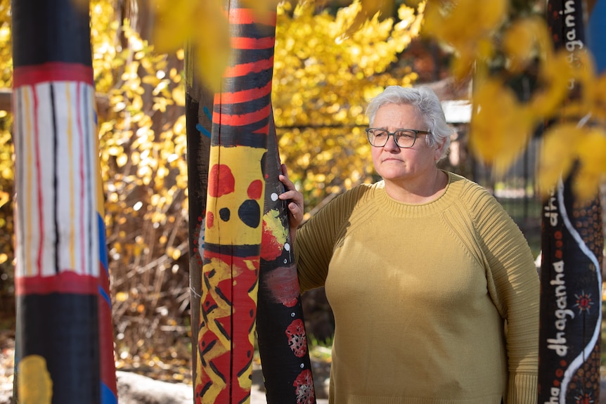 A woman standing amongst Indigenous art, yellow leaves of trees surrounding her. 