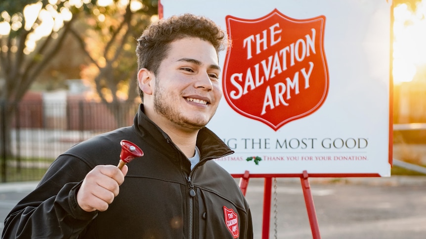 Man shaking bell, standing in front of The Salvation Army sign, raising funds.