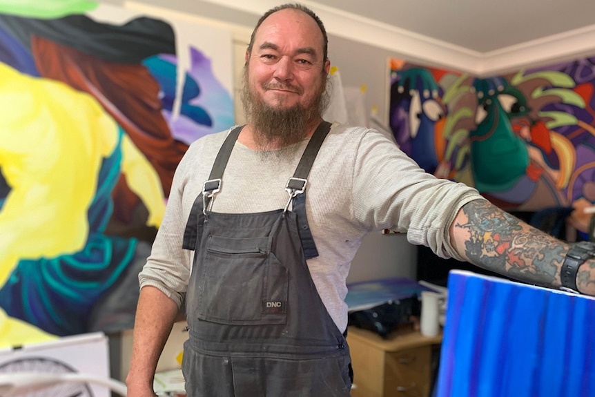 Mid shot of artist Russell Sanderson standing in a room, with canvas paintings in background