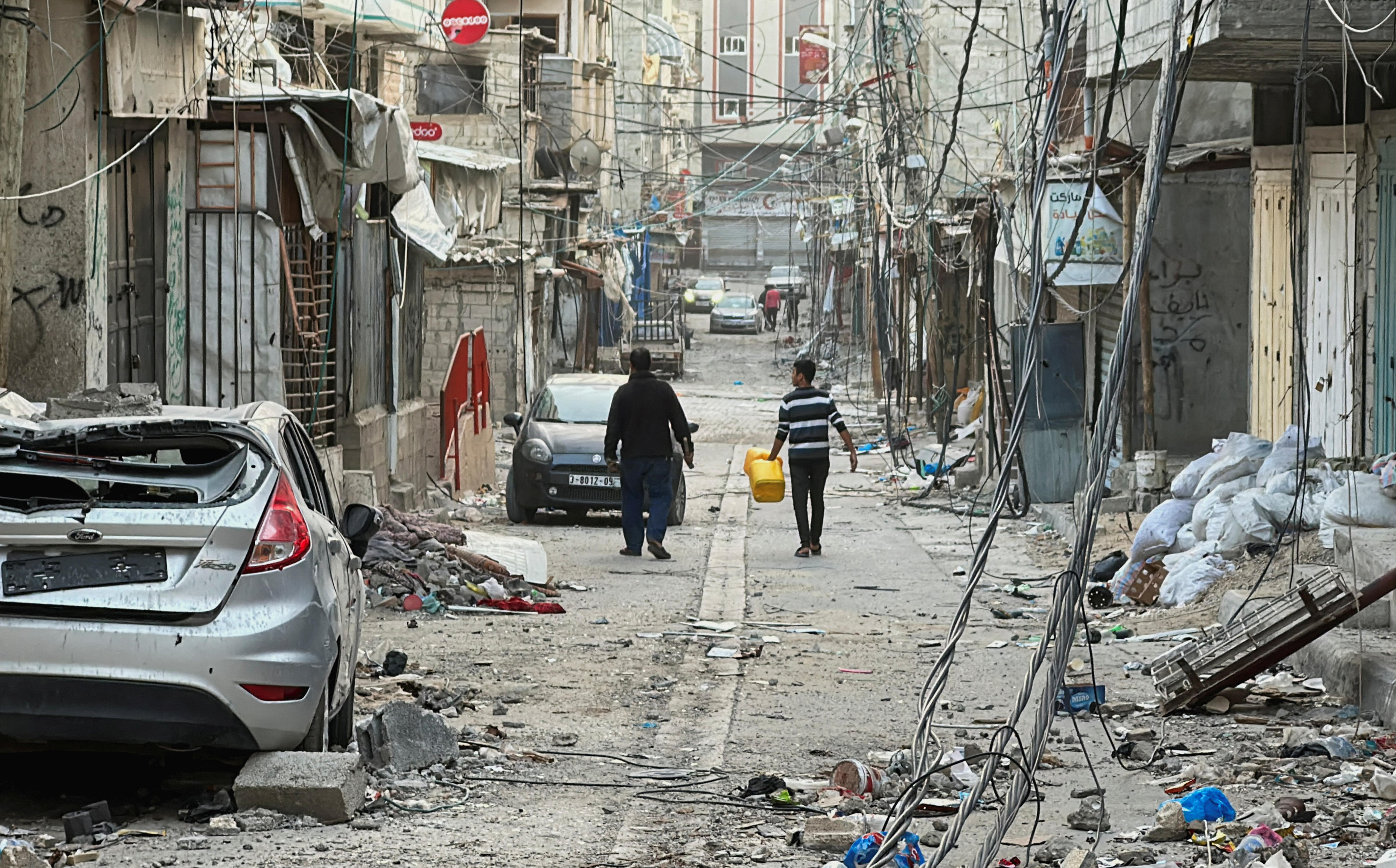 A wide shot of a damaged road, with a damaged car and rubble on it. Two men walk down the middle of the street, away from camera