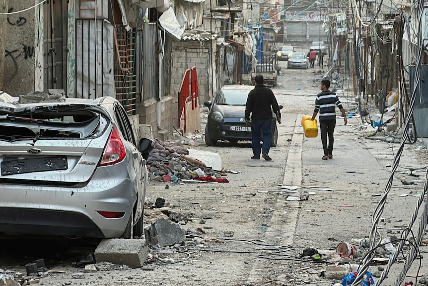 A wide shot of a damaged road, with a damaged car and rubble on it. Two men walk down the middle of the street, away from camera