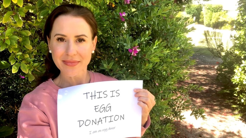 A woman stands in front of a bush holding a sign that says "This is egg donation"
