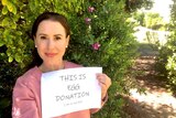 A woman stands in front of a bush holding a sign that says "This is egg donation"