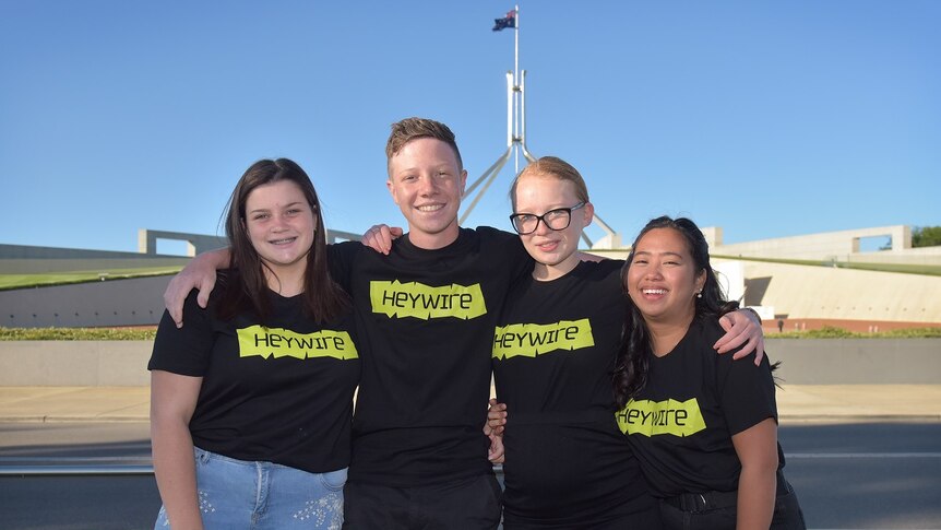 Three young women and a young man in Heywire shirts stand in front of Parliament House with arms around each other.