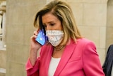 Nancy Pelosi wears a pink jacket and a facemask as she speaks on her phone, security are behind her.