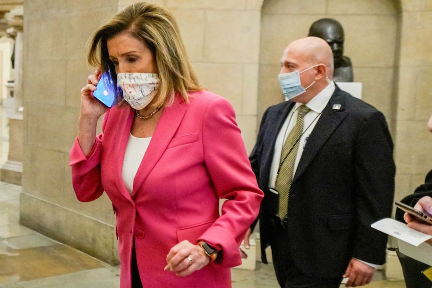 Nancy Pelosi wears a pink jacket and a facemask as she speaks on her phone, security are behind her.