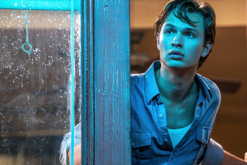 Young white man with windswept hair and blue shirt looks with a sense of wonder past a window illuminated in vivid cyan light.