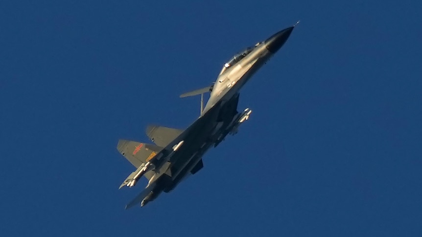 A green and grey military jet flies through clear blue sky.