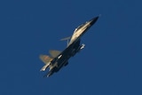 A green and grey military jet flies through clear blue sky.