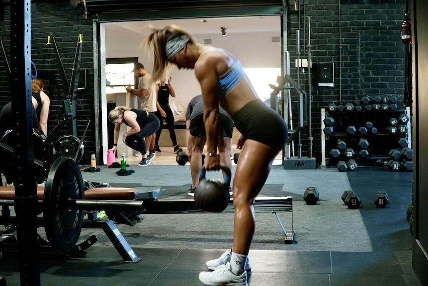 A woman exercises with a kettle bell in a gym, she has long blonde hair and a hairband