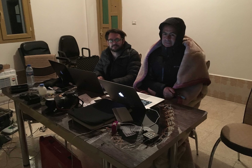 Hollett and Brown wearing coats and blankets sitting in front of lap tops on desk in room.