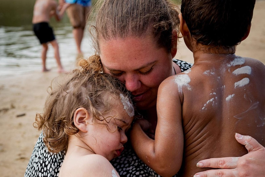 Two young Indigenous children with clay paint on their bodies are embraced by their mother