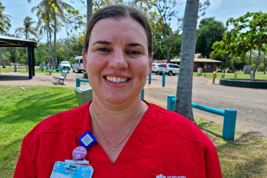 A woman in a red shirt smiling at the camera, with a park in the background. 