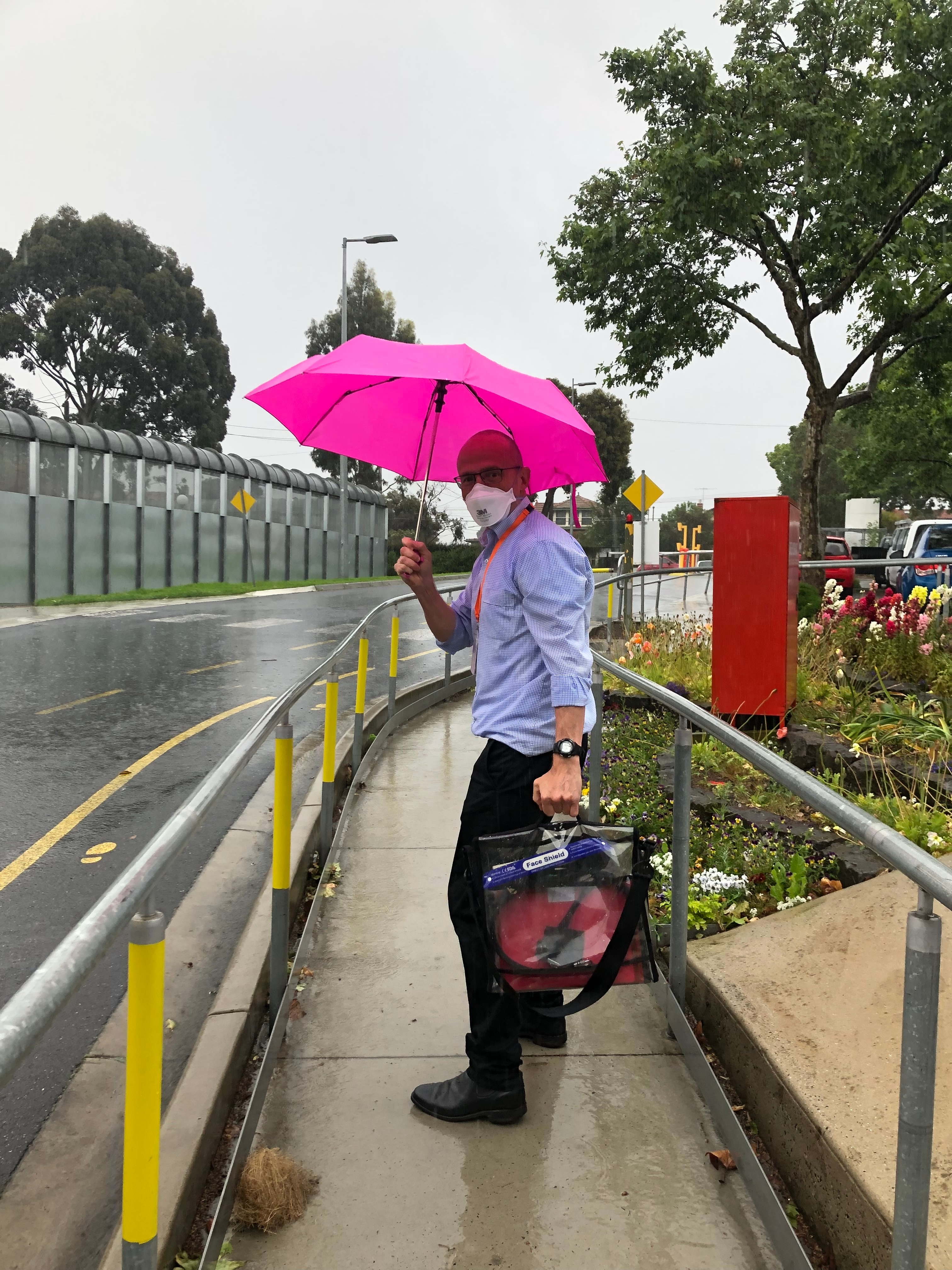 A man wearing a facemask, holding a bright pink umbrella and a clear briefcase, standing on a footpath outside a prison.