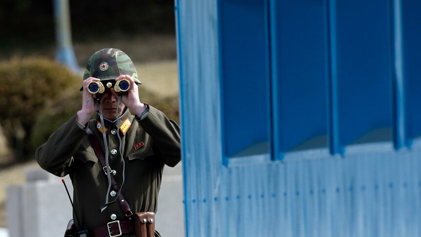 A man dressed in an army uniform looks through gold binoculars next to a blue building.