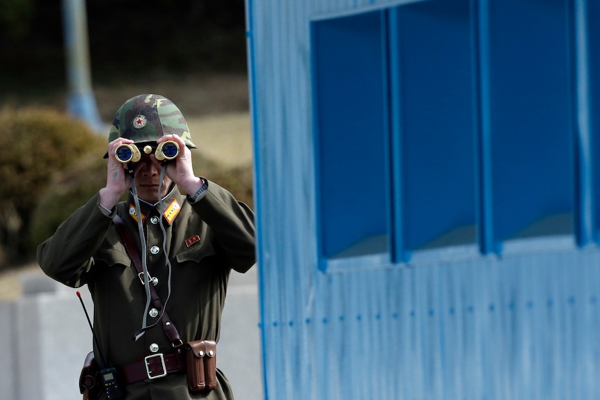 A man dressed in an army uniform looks through gold binoculars next to a blue building.