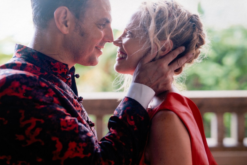 Caucasian man holds his wife's face in his hands. They are looking into each other's eyes and smiling.
