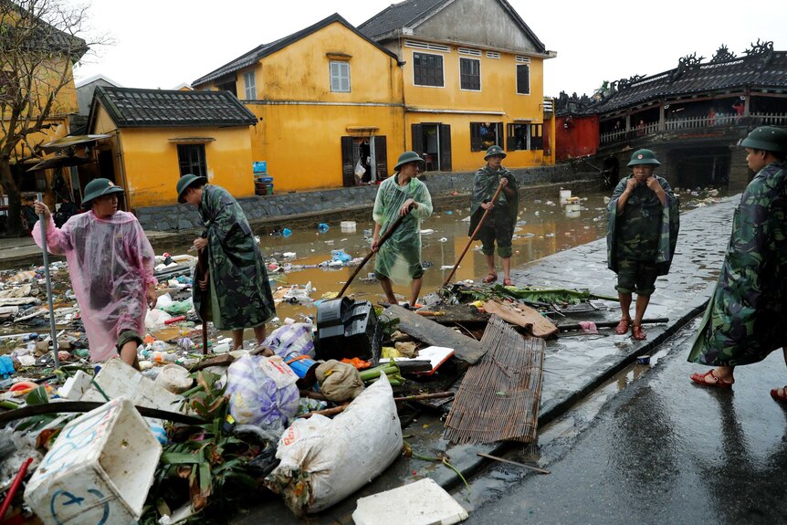 Vietnam soldiers stand among rubbish wearing ponchos and holding brooms in flooded streets in Hoi An.
