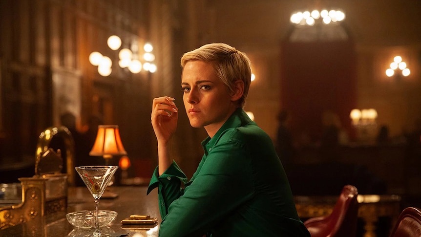 A woman with short blonde hair in emerald green shirt rests on ledge of bar and sits on bar tool in warmly lit room.