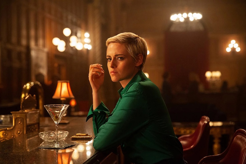 A woman with short blonde hair in emerald green shirt rests on ledge of bar and sits on bar tool in warmly lit room.