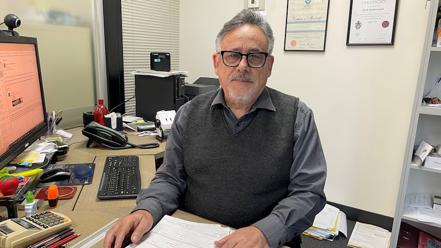 A man with grey hair and glasses sits at a desk in his office.