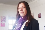 A woman with long brown hair stands in a lounge room wearing a black blazer, white shirt and purple scarf.