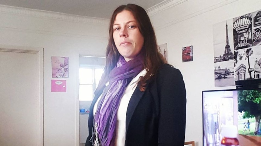 A woman with long brown hair stands in a lounge room wearing a black blazer, white shirt and purple scarf.