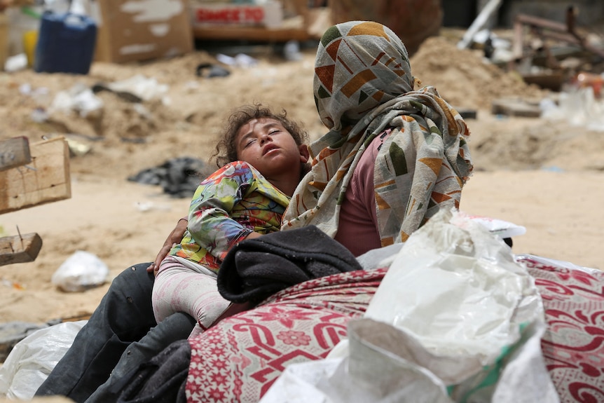 A woman in a veil holds a young child sleeping in her arms surrounded by debris in bright sunshine
