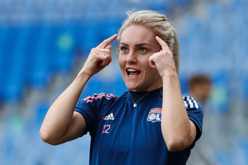 Ellie Carpenter points to her head with both hands, wearing a dark blue t-shirt