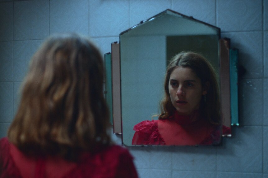 A 25-year-old woman with brown shoulder-length hair, in a fancy red top, staring into a mirror
