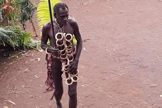 A chief from Vanuatu wearing a necklace of pig tusks walking with a wooden spear in front of a group of women in grass skirts.