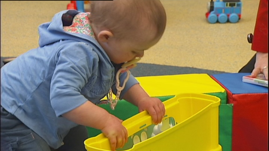 Video still: Good generic photo of a young toddler playing with a bright yellow toy at child care. Aug 2012