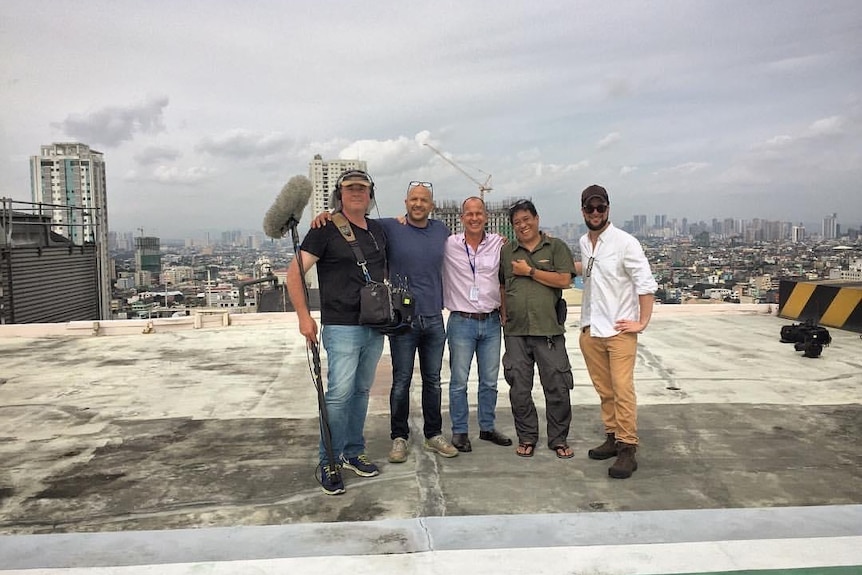 Five men standing on a rooftop with arms around each other and one man holding microphone on a boom pole.