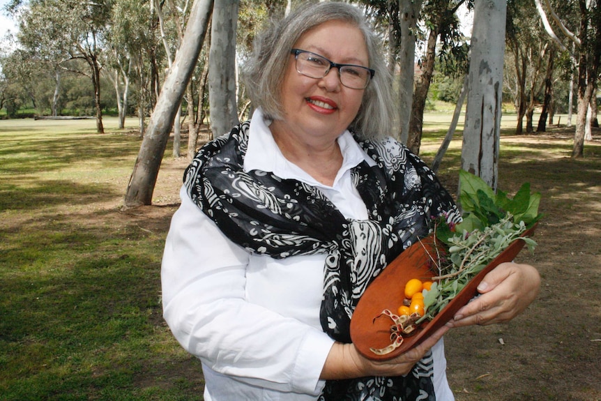 Dale Tilbrook holds a traditional wooden oblong bowl filled with indigenous cooking ingredients.
