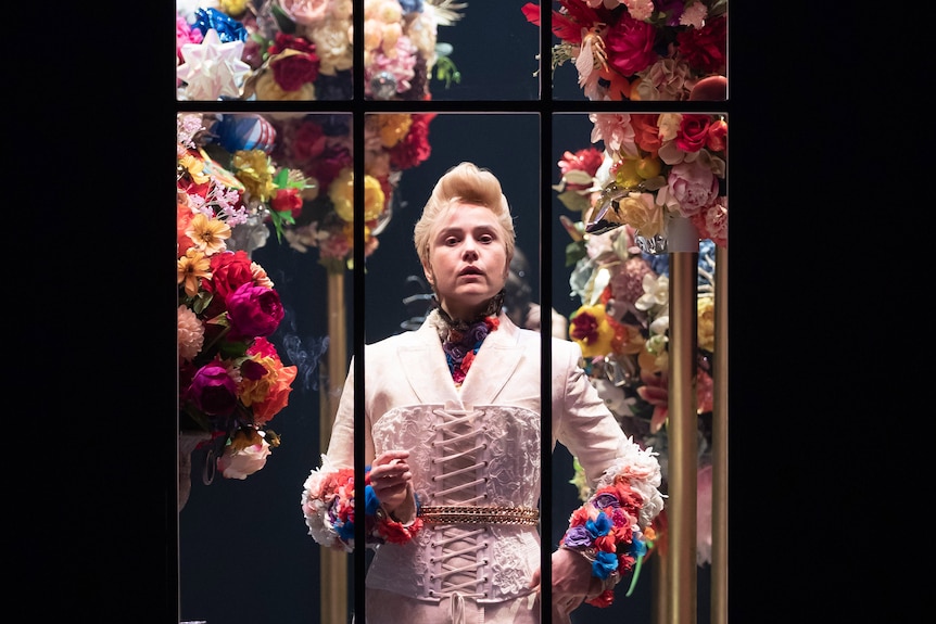 White woman dressed as man with blonde quaffed hair and regal white suit stands among floral bouquets behind glass window