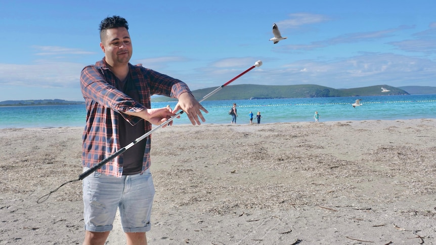 Man twirls white cane like a baton on the beach as a seagull flies past.