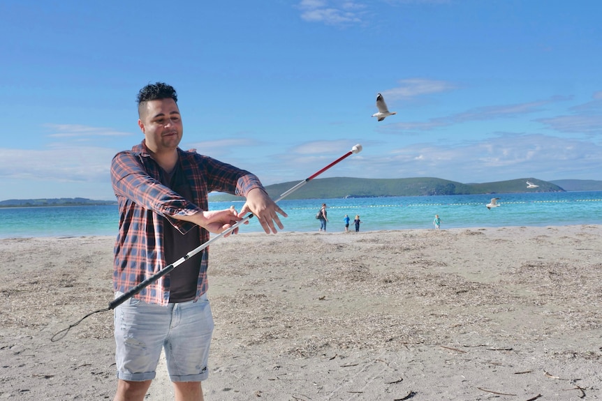 Man twirls white cane like a baton on the beach as a seagull flies past.