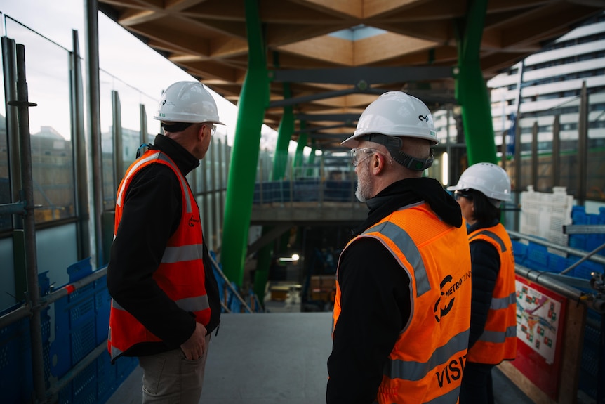 Three people in hard hats and high-vis at the top of a stairwell with a wooden structure overhead.