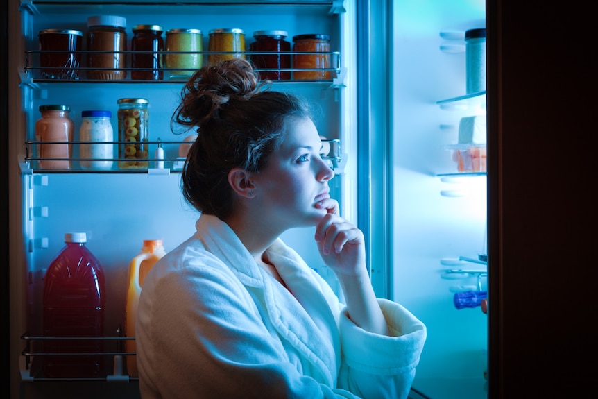 A young white woman in a bathrobe looking into an open fridge
