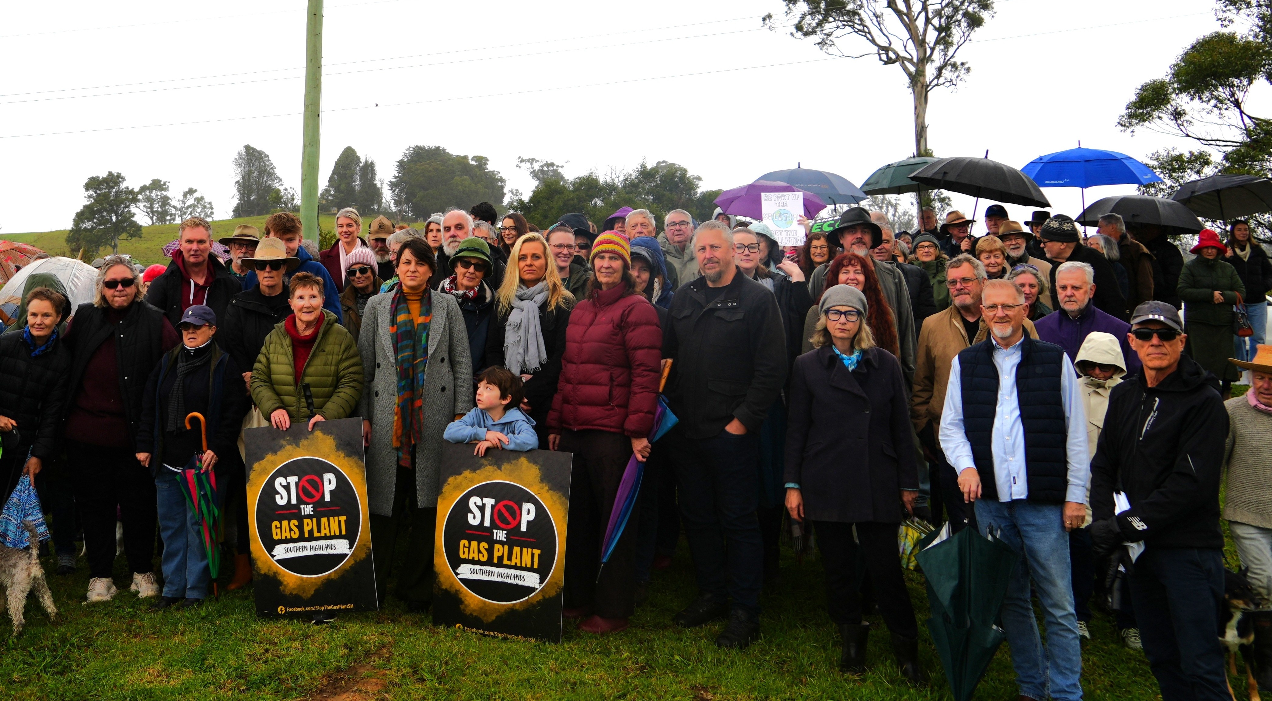 large crowd of people holding 'stop the gas plant' signs on a hill