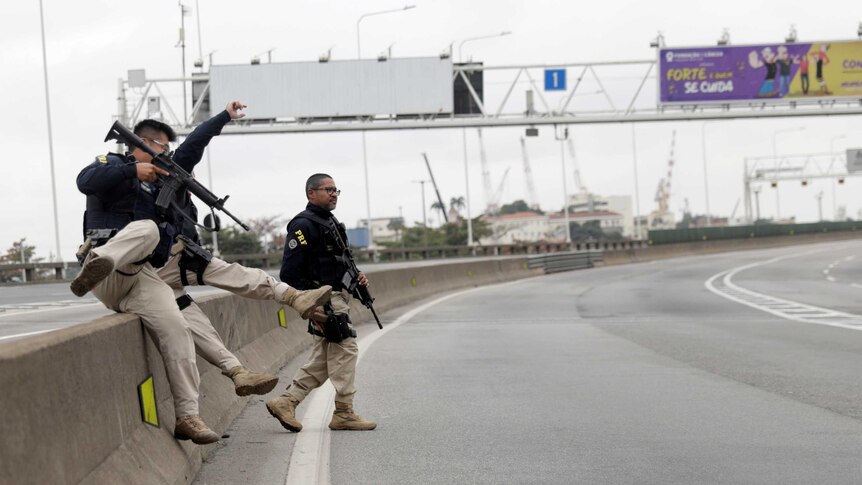 Police with guns jump over a road barrier.