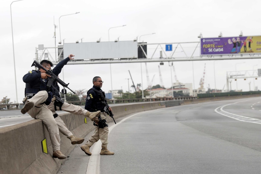 Police with guns jump over a road barrier.