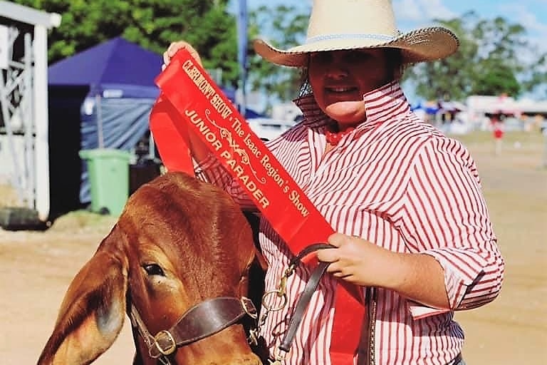 A young lady in a white and red striped shirt and jeans holding a red winner's ribbon stands next to a brown cow.