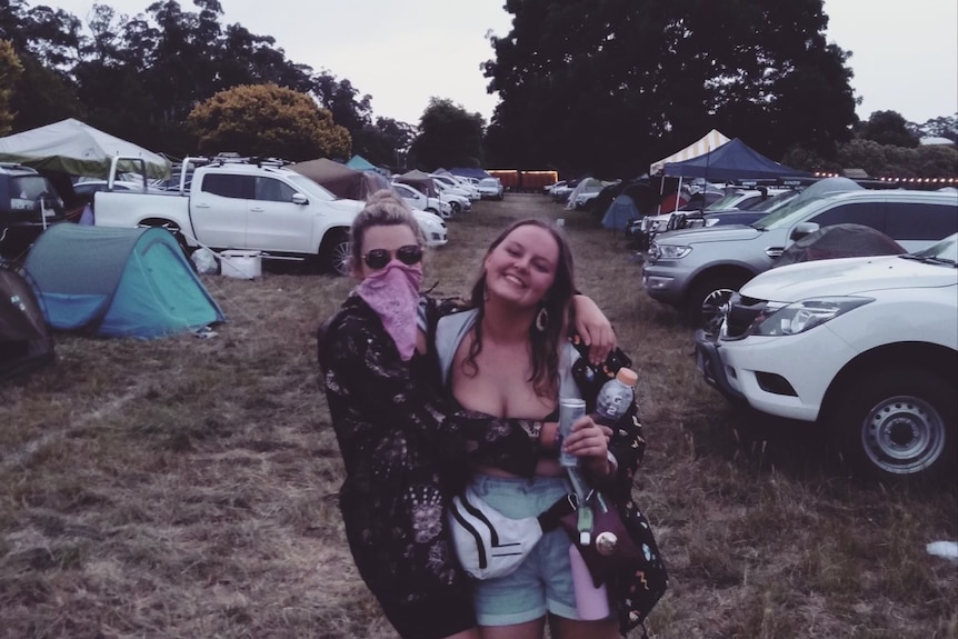 Two girls hug each other standing in a campground at a music festival.