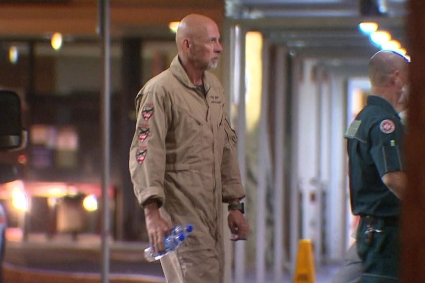 A man in a light brown flight suit walks while carrying a water bottle