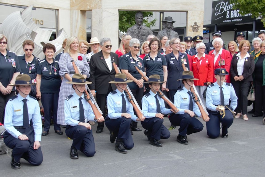 former women personnel of armed forces stand in a group behind young women army cadets who are kneeling on the ground