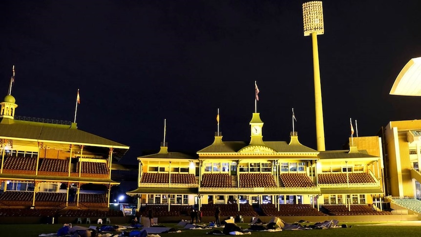 A group of people sleep in makeshift cardboard shelters in front of the members stand at the SCG