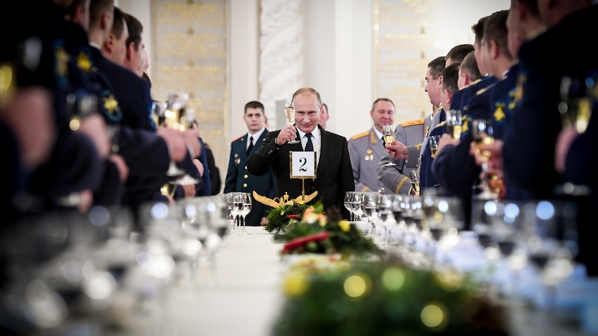 Vladimir Putin holding a champagne flute aloft in front of a table lined with standing Russian soldiers