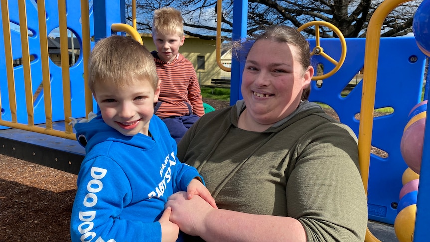 A woman and two boys sitting in a playground smile at the camera.